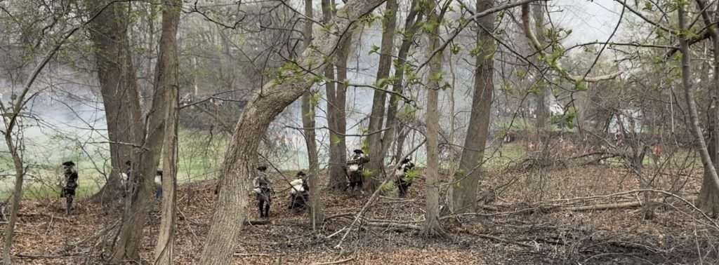 Group of people in historical clothing standing in a leaf-barren forest with smoke in the distance.