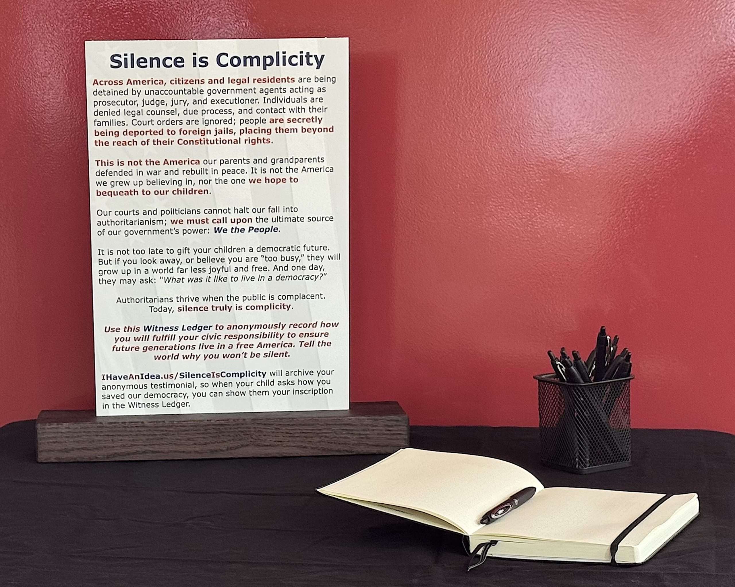 table ledger pen placard ‣ I Have An Idea Poster titled 'Silence is Complicity' on a wooden table, with a notebook and pen in foreground and a pencil cup against a red wall backdrop.