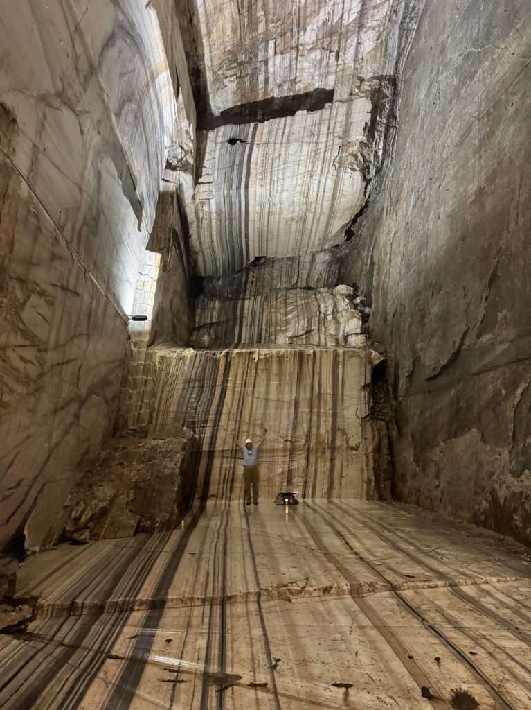 Person with arms raised stands at the base of a large, vertical quarry or mine shaft with striated rock walls and rails on the floor for scale and access.