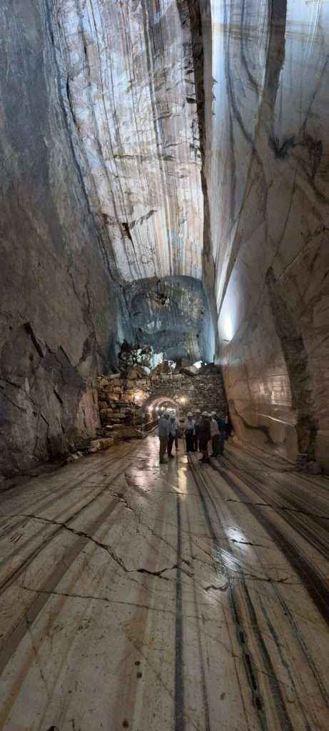 Inside a deep stone quarry shaft with striped rock walls and a group of workers wearing helmets near a stone cairn, lit by artificial lights.