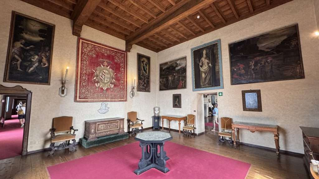 Historic interior gallery with large framed paintings on beige walls, red carpet, and heavy wooden ceiling beams.