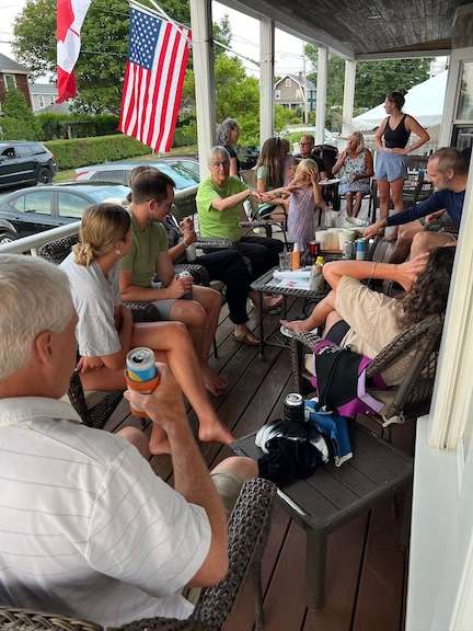 Front Porch ‣ I Have An Idea Group of people sitting on a porch around a table, chatting with drinks; American and Canadian flags hang nearby.