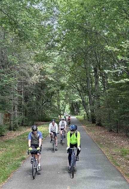 IMG_1088 1 ‣ I Have An Idea Six cyclists wearing helmets ride along a tree-lined paved path through a forest.