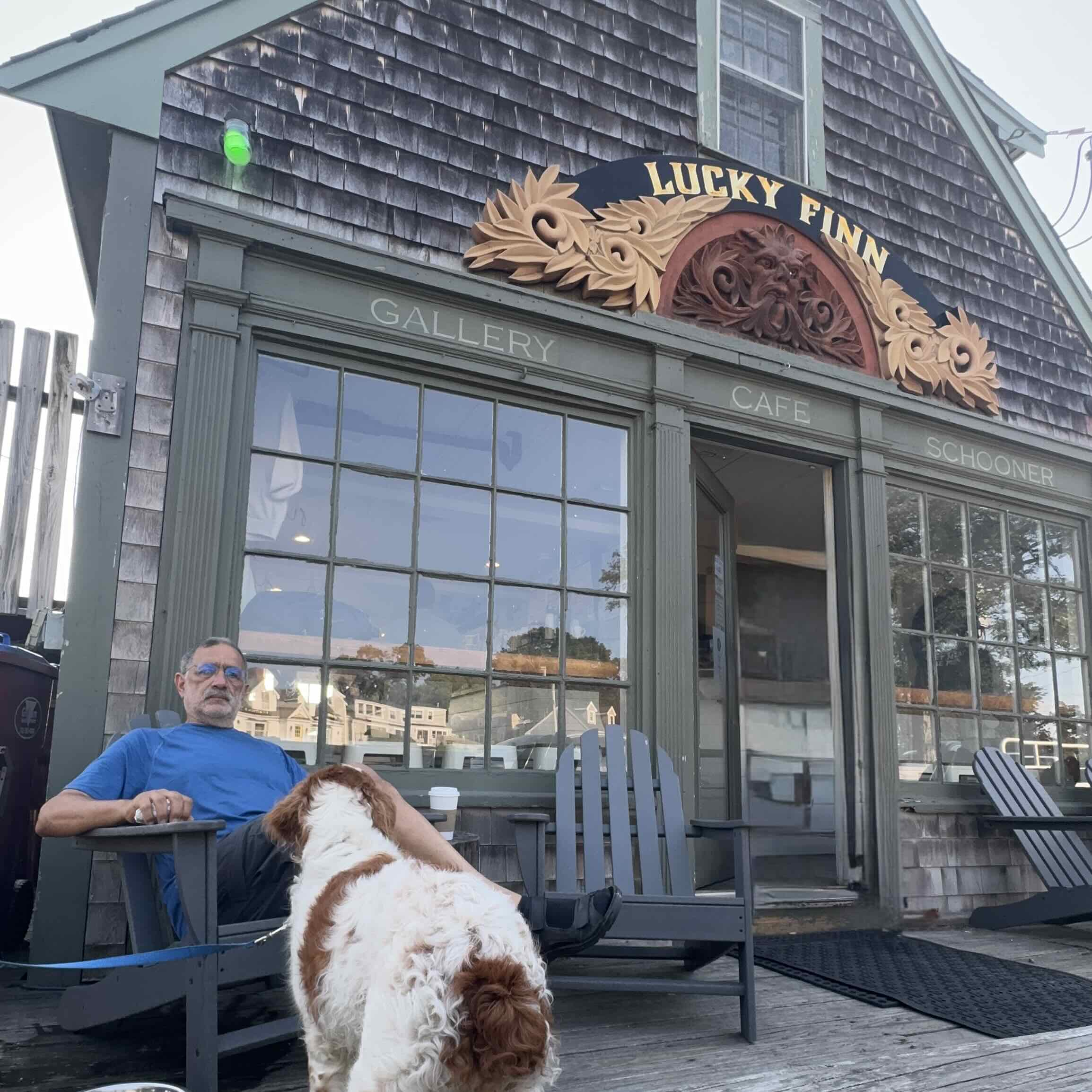 Man in a blue shirt sits at an outdoor cafe table with a white and brown dog in front of a gray storefront with a decorative sign reading'Lucky Finn' and 'Gallery' above the windows.