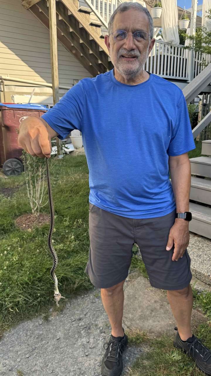 IMG_1294 1 ‣ I Have An Idea Older man in a bright blue t-shirt and gray shorts holds a long snake by the tail outdoors in a backyard setting.