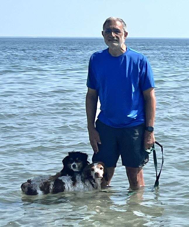 IMG_4856 ‣ I Have An Idea Man wearing a bright blue shirt stands in shallow ocean water with two dogs at his feet.