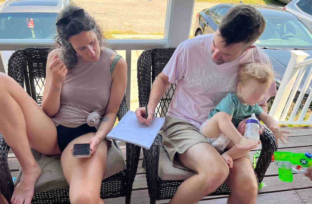 Kim and Daniel2 ‣ I Have An Idea Family on a porch chair: a woman with phone in hand and a man holding a toddler who drinks from a bottle while another person writes on a clipboard.