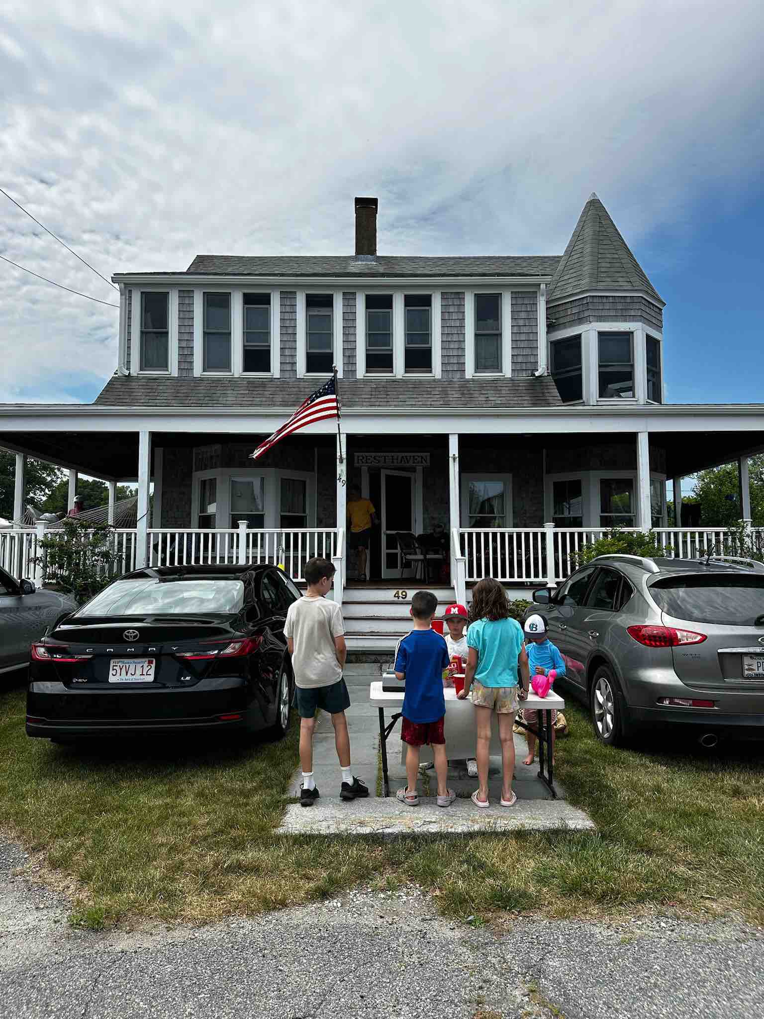 Rest-Haven ‣ I Have An Idea Front view of a gray two-story house with a turret, an American flag, and kids gathered around a lemonade stand on the porch area.