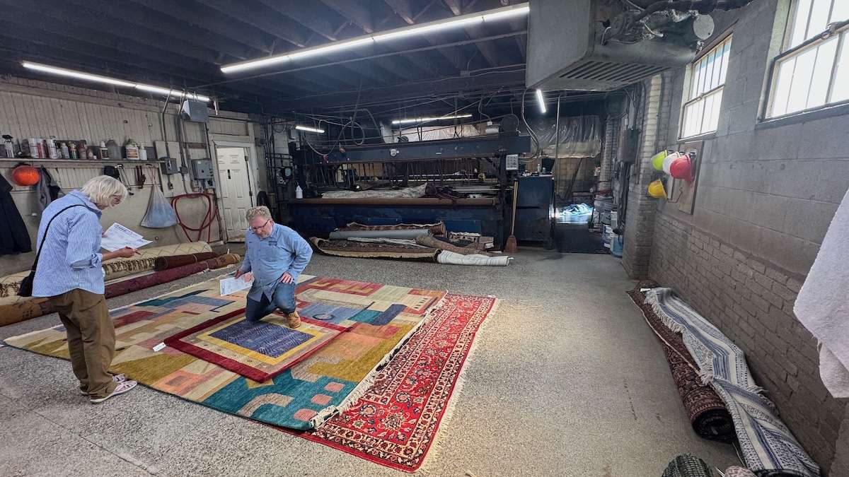 IMG_1905 ‣ I Have An Idea Two men examine stacked, multicolored rugs laid on a workshop floor, with more rolls and machinery in the background.