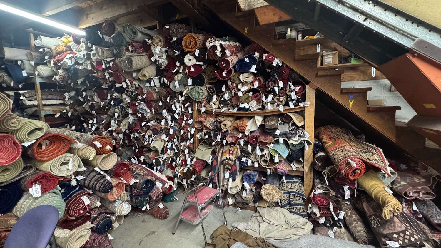 IMG_1910 ‣ I Have An Idea Cluttered rug shop storage with dozens of rolled carpets stacked on shelves under a wooden staircase; a red step ladder in front.