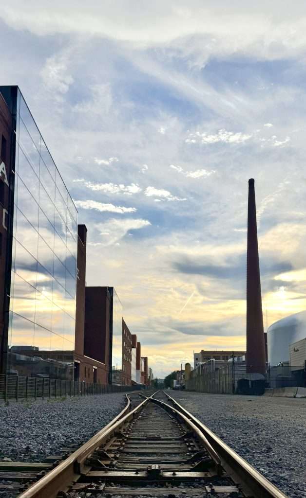 Railroad tracks lead into an industrial area with a glass-fronted building on the left and a tall smokestack on the right under a cloudy sky at sunset.